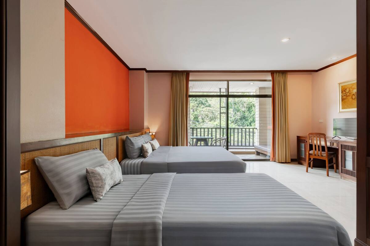 A hotel room with two beds, a desk, and a balcony with a view of greenery.
