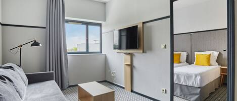 A modern hotel room with a sofa, a small wooden table, a TV mounted on the wall, and two beds with yellow pillows.
