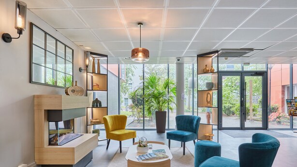 Un vestíbulo moderno con un mostrador de recepción de madera, sillas azules y amarillas, una mesa de centro y ventanas amplias con vistas al exterior.