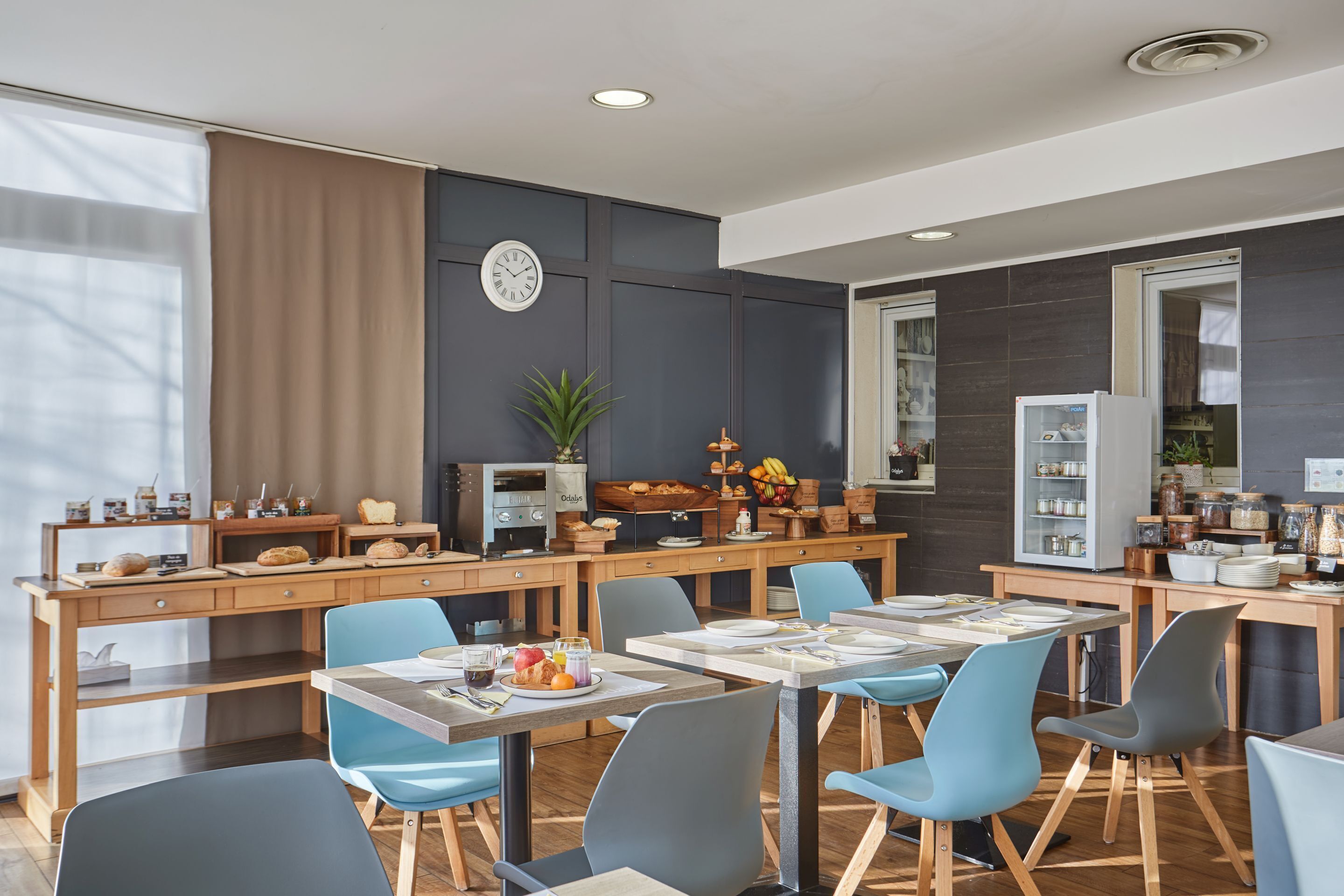 A modern dining area with wooden tables and blue chairs, a clock on the wall, and a variety of food items displayed on shelves.