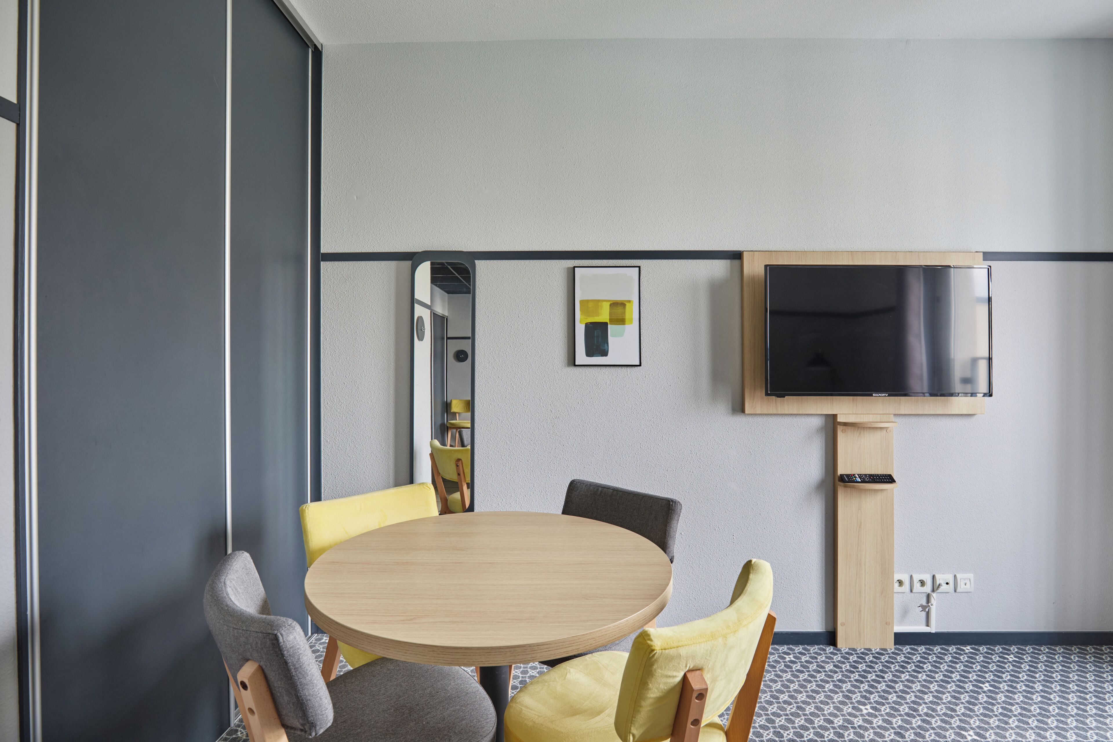 A modern meeting room with a round table, four chairs, a flat-screen TV mounted on the wall, and a small framed artwork above the TV.