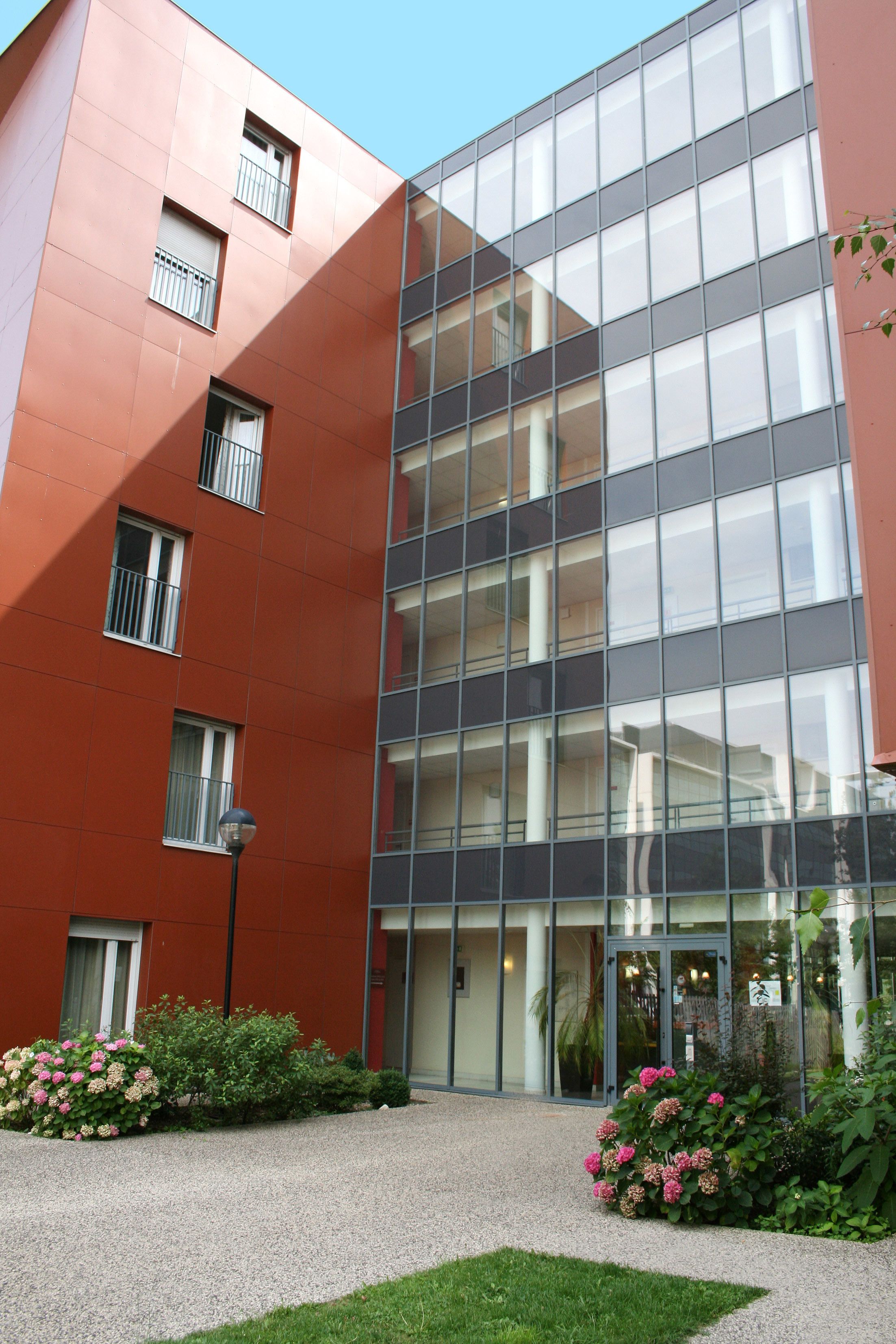 A modern building with a glass facade, multiple balconies, and a gravel courtyard with pink flowers.