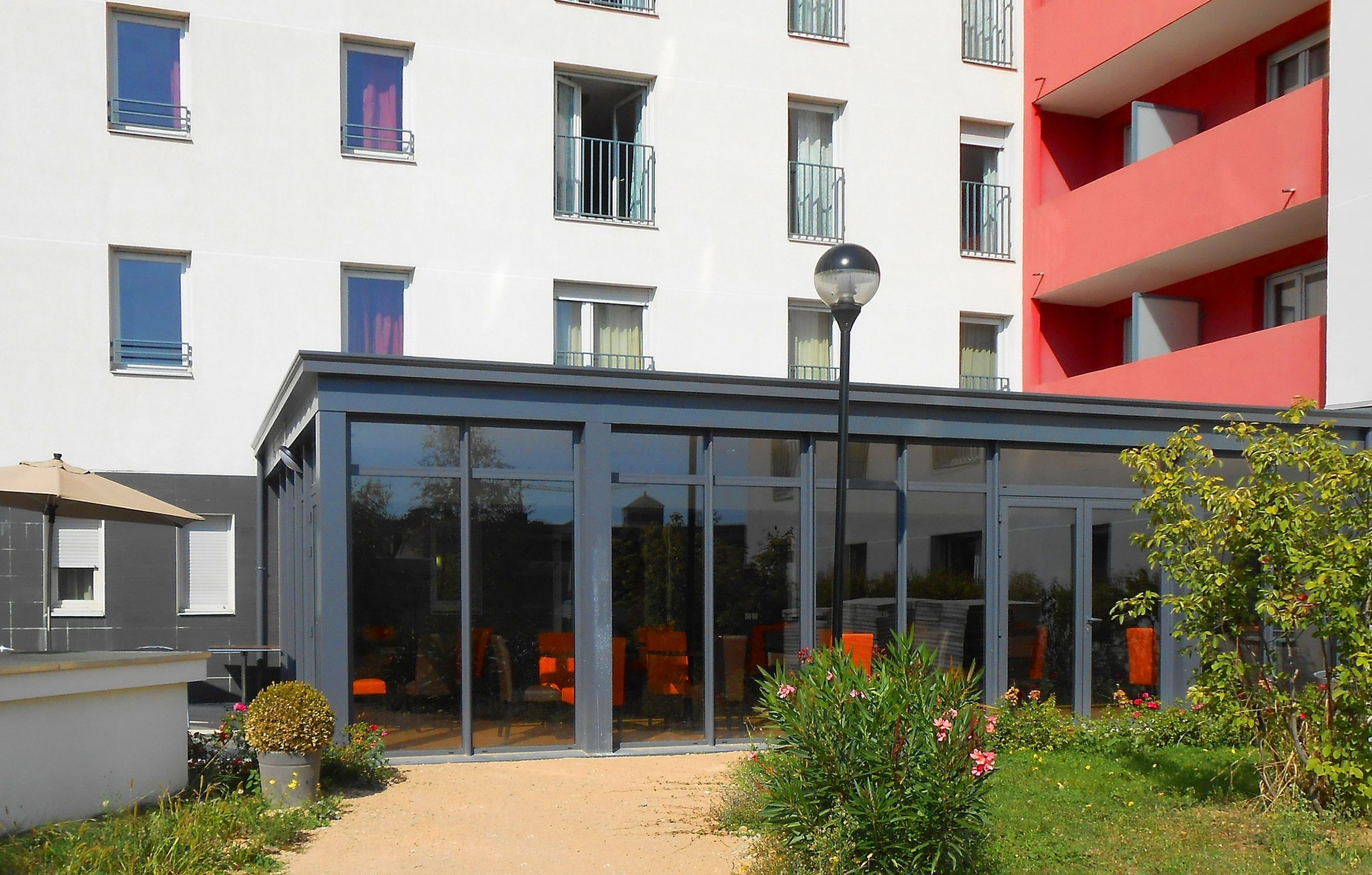 Un edificio moderno con un invernadero de vidrio, asientos exteriores y un balcón rojo.