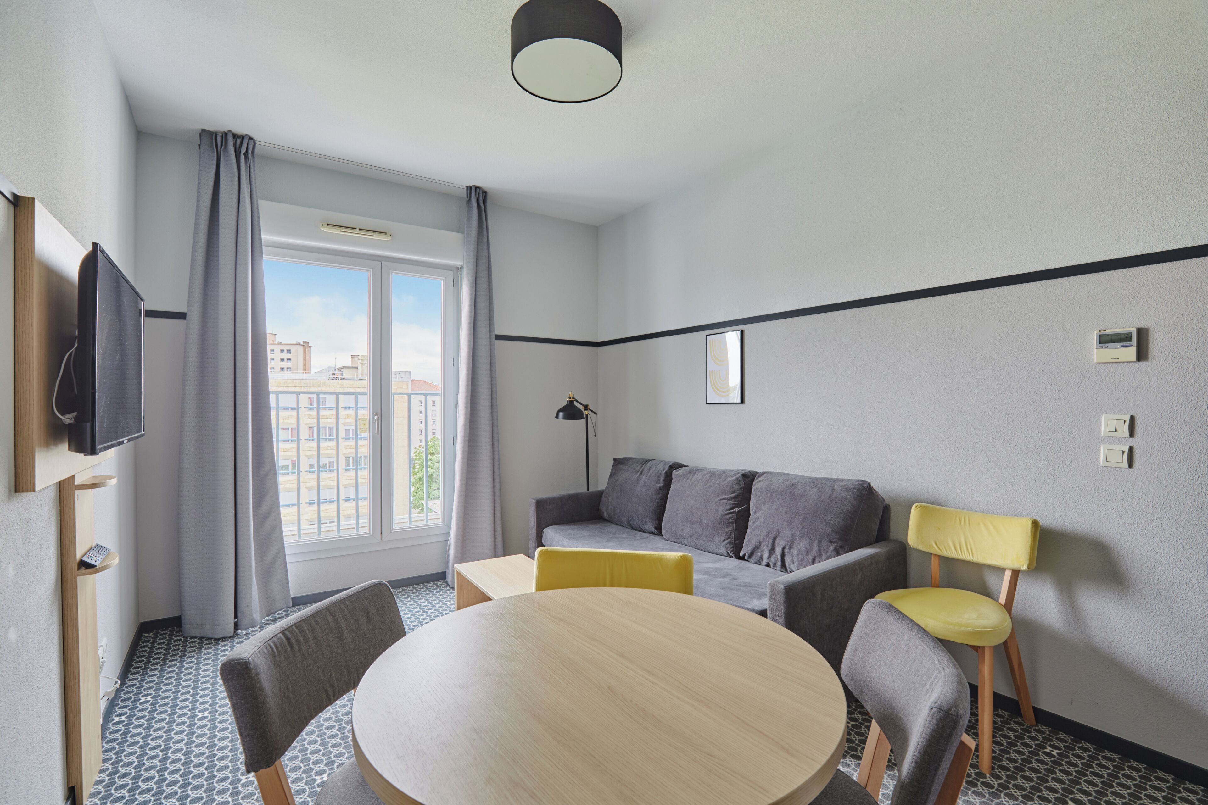 Habitación de hotel con una mesa de comedor redonda, dos sillas, un sofá, un televisor montado en la pared y una ventana con cortinas.