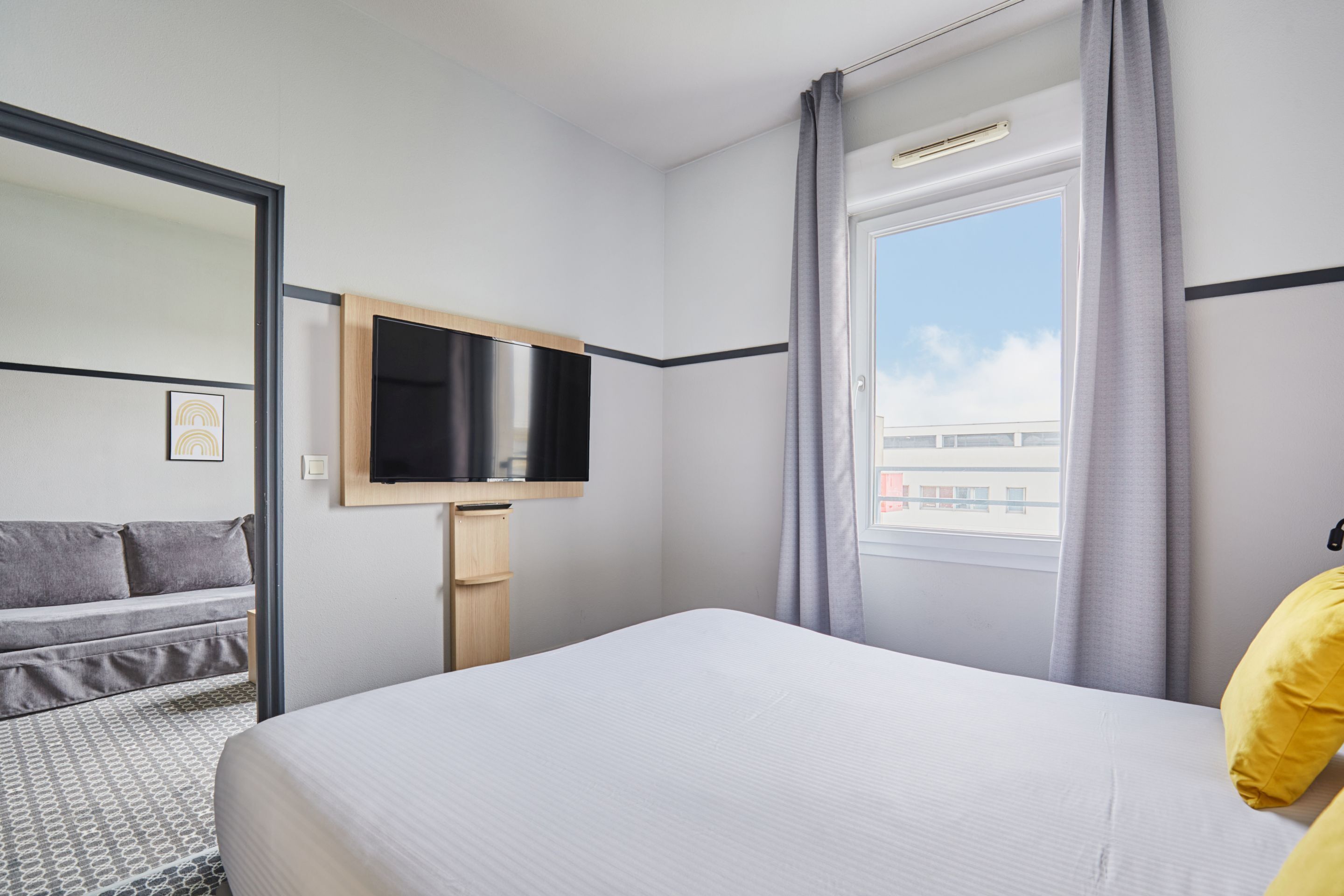 Habitación de hotel con cama, televisor montado en la pared, ventana con cortinas y sofá con alfombra estampada.