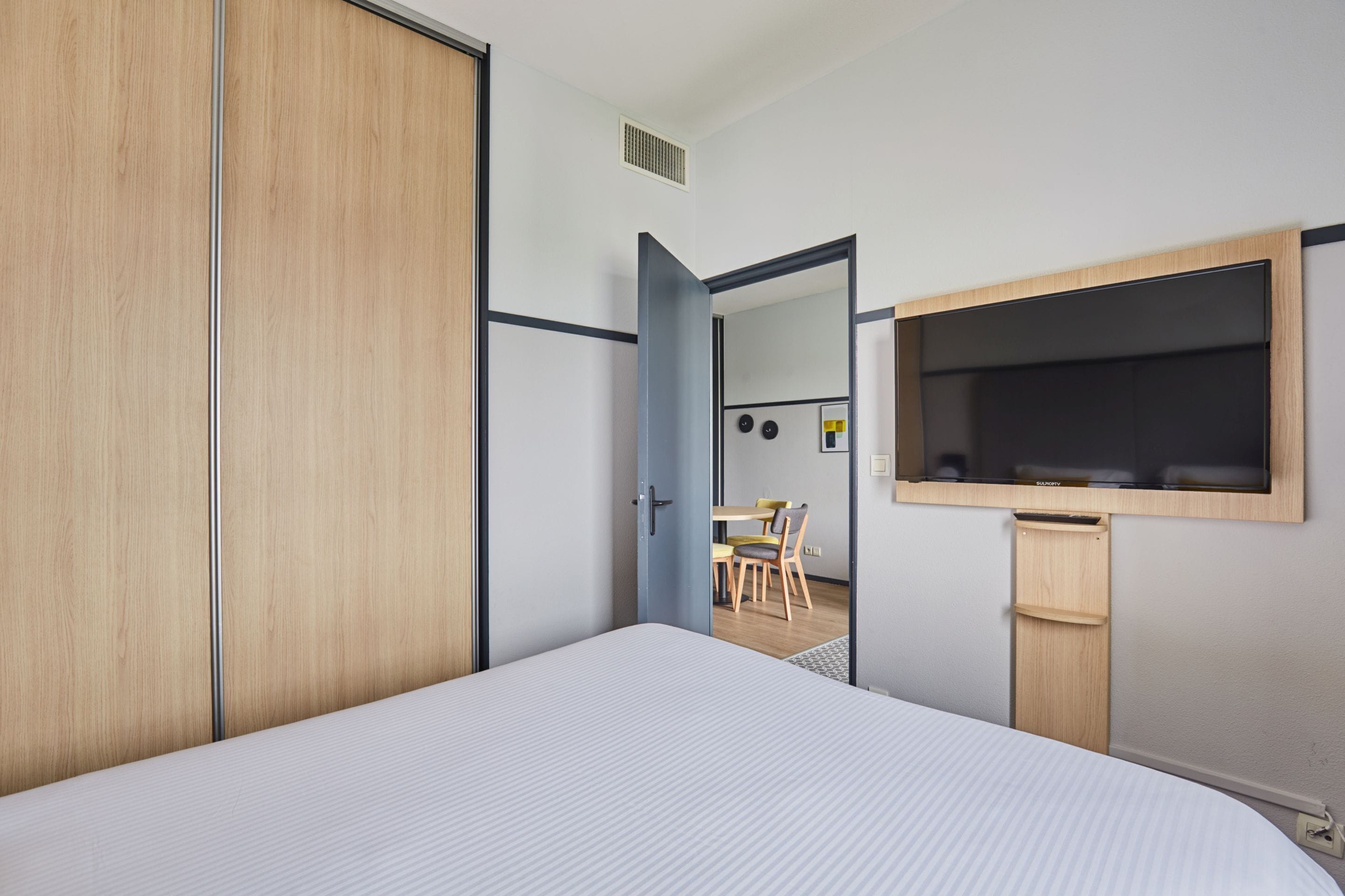 Een slaapkamer met een bed, een houten kledingkast, een aan de muur bevestigde televisie en een kleine tafel met stoelen op de achtergrond.