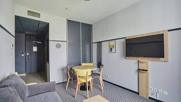 Een hotelkamer met een bank, een eettafel met stoelen, een aan de muur bevestigde televisie en een deur naar een andere kamer.