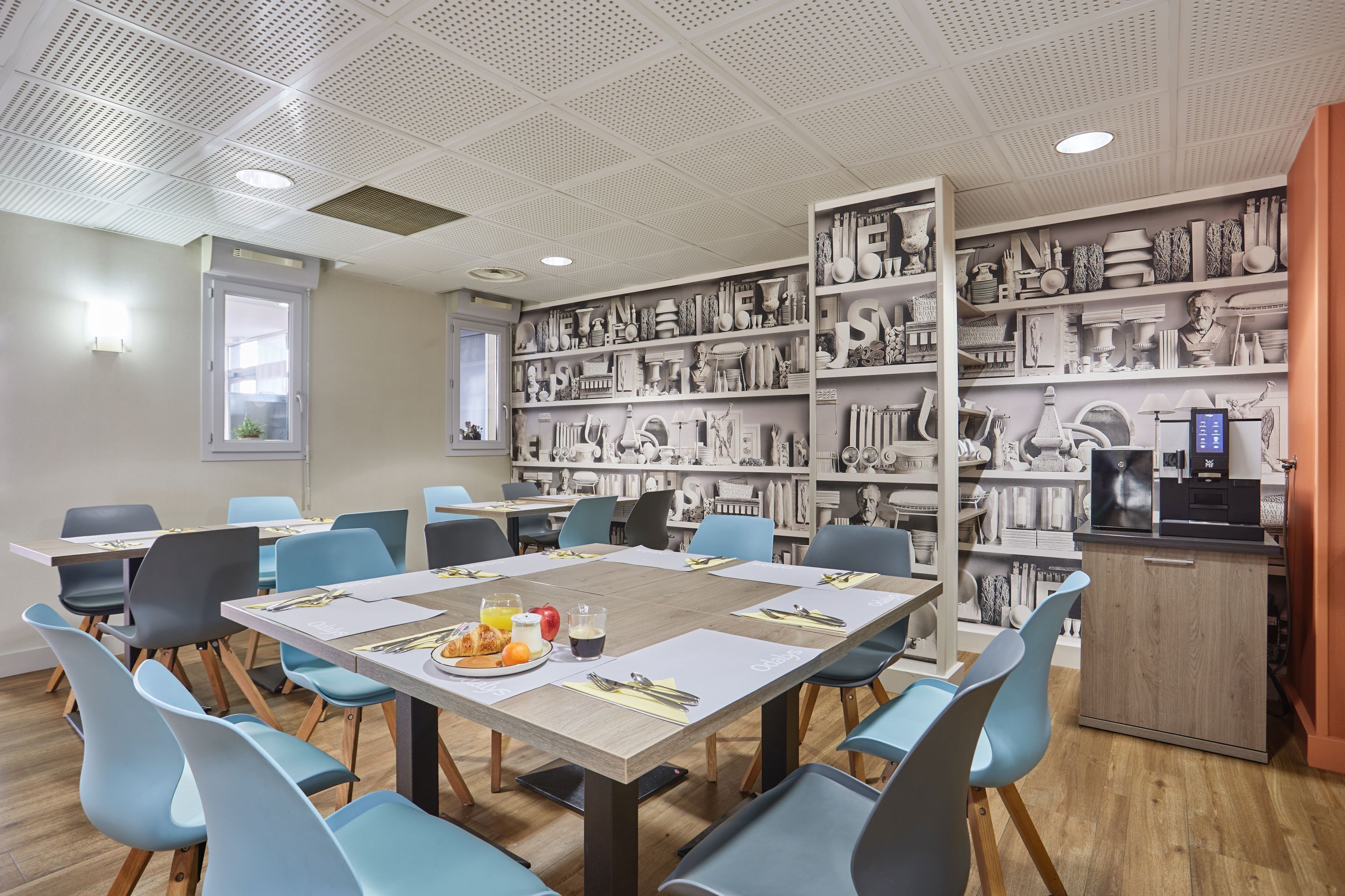 Un comedor con mesas dispuestas para cuatro, sillas y una pared decorada con objetos.