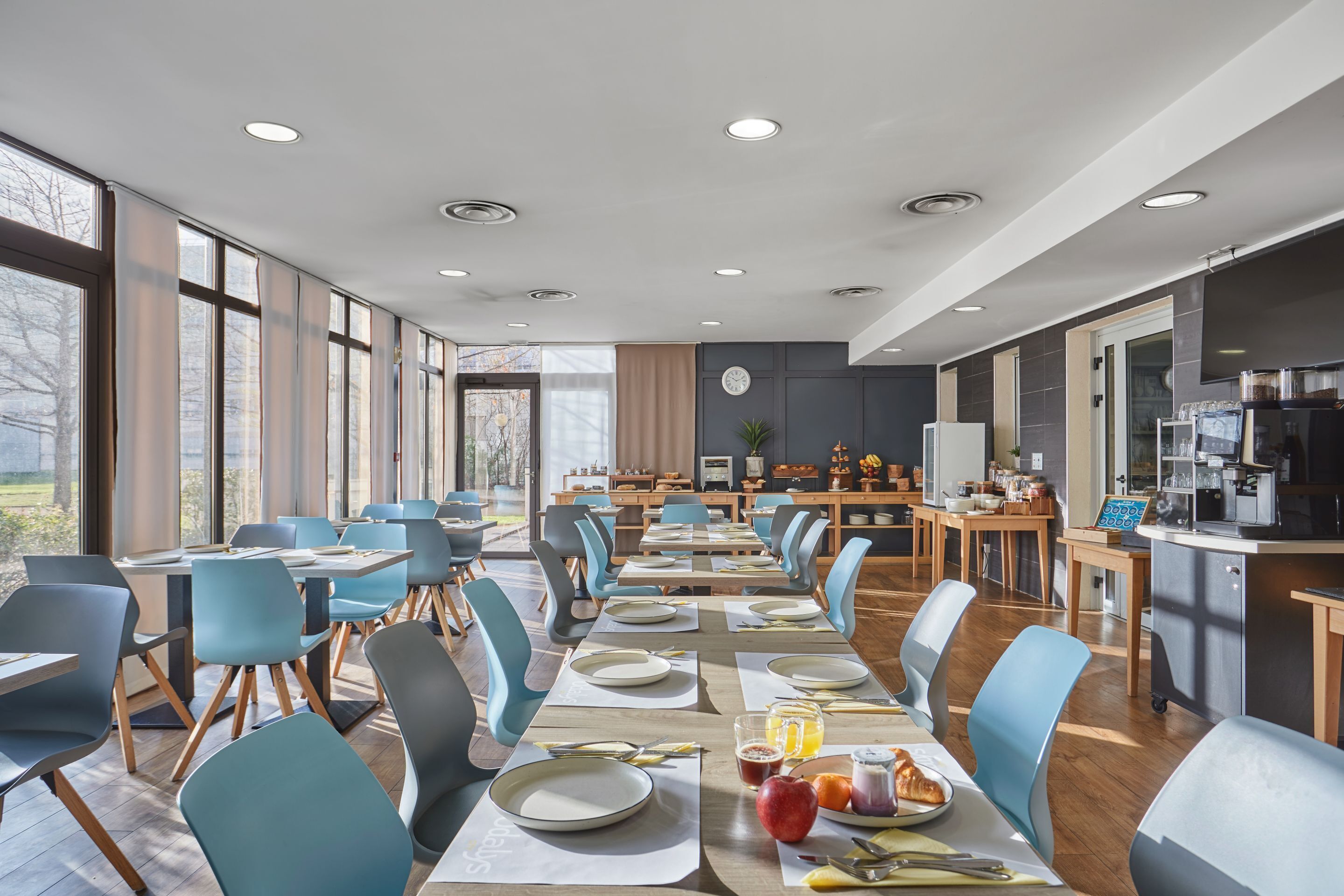 Un comedor moderno con mesas largas, sillas azules y una cocina al fondo.