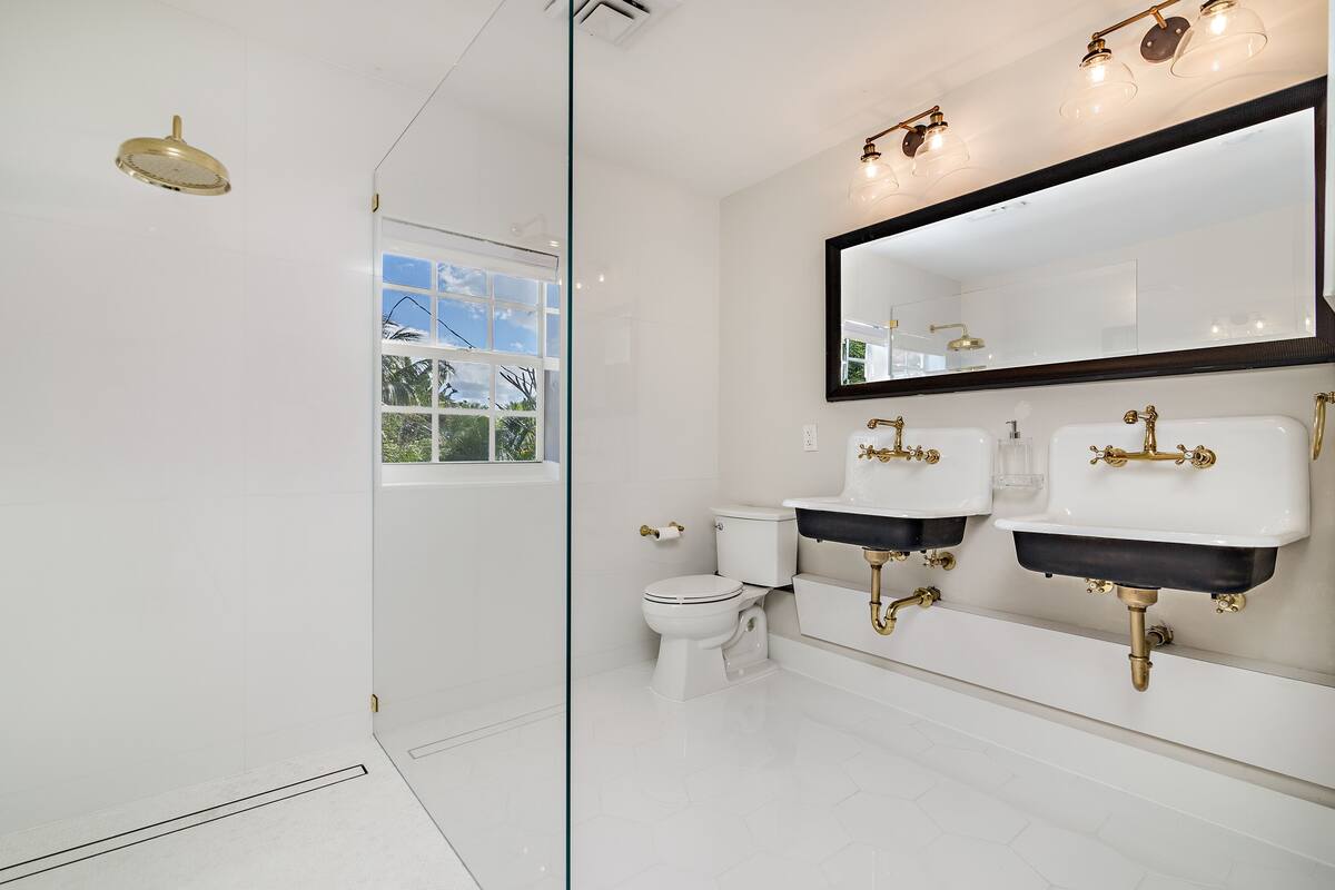 Une salle de bain moderne dotée d’une douche à parois de verre, de toilettes et de deux lavabos avec des robinetteries dorées.