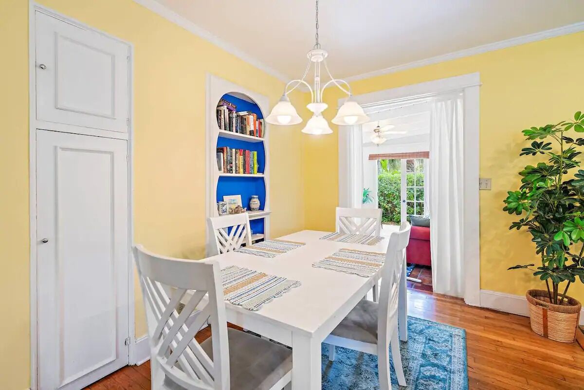 A dining area with a white table, chairs, and a blue rug. There is a bookshelf with books and a plant in the corner.