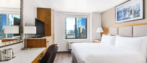 Quarto de hotel com cama, escrivaninha com computador, cadeira, janela grande com vista para a cidade e quadro na parede.