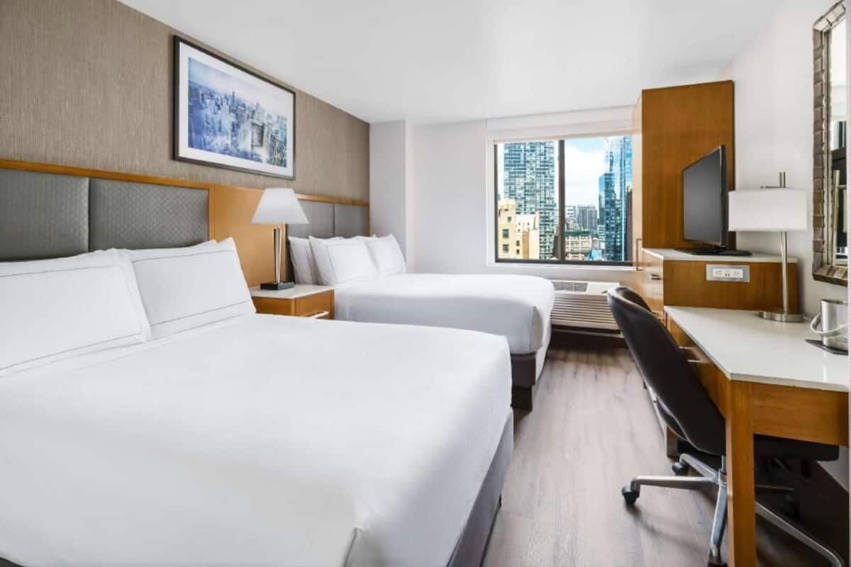 A hotel room with two beds, a desk, a chair, a TV, and a window with a city view.