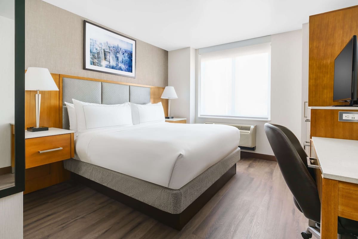 A hotel room with a large bed, two bedside tables with lamps, a flat-screen TV, and a window with blinds.