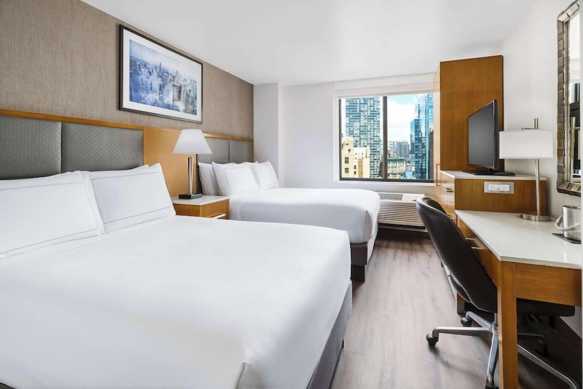 Quarto de hotel com duas camas, uma escrivaninha com computador, televisão e janela com vista para a cidade.