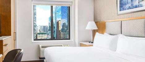 A hotel room with a large bed, a desk, a chair, a window with a city view, and a framed picture on the wall.
