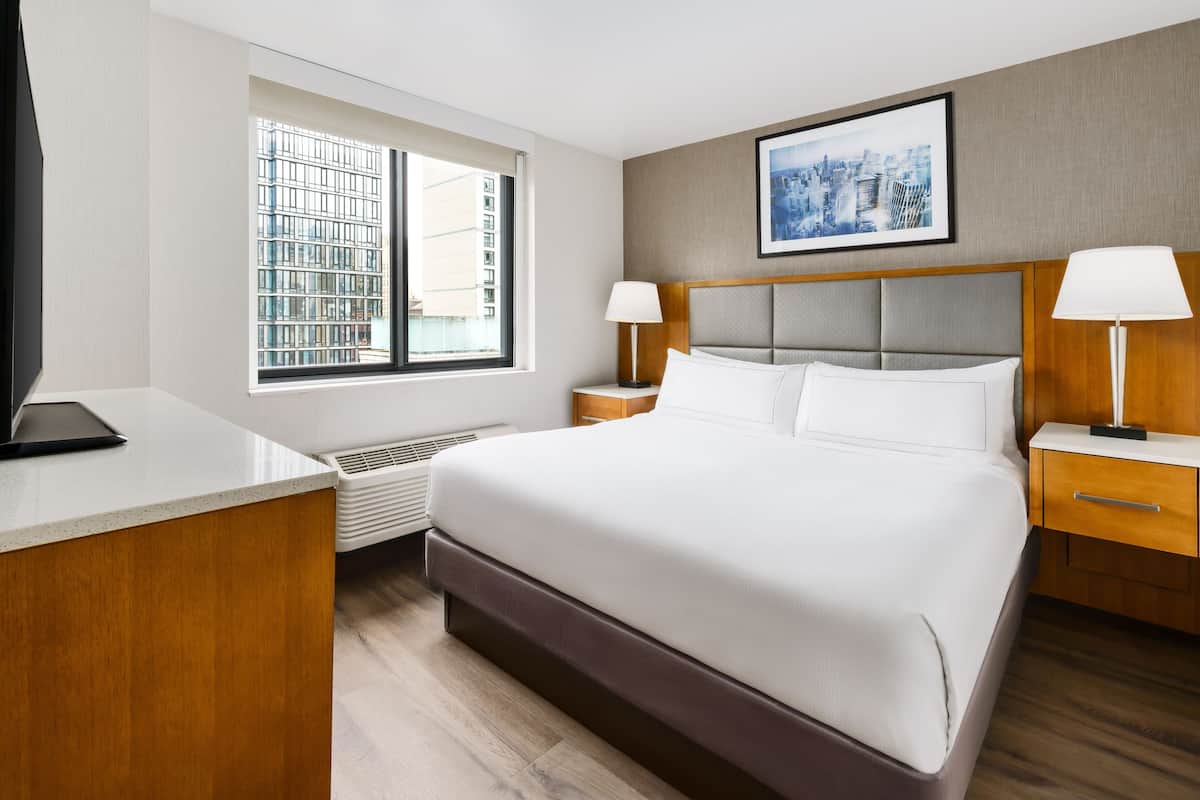 A hotel room with a large bed, two bedside tables with lamps, a window with a city view, and a framed picture on the wall.