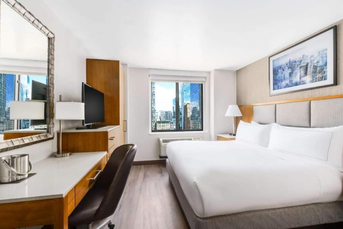 A hotel room with a bed, a desk, a chair, a television, a window with a city view, and a framed picture on the wall.