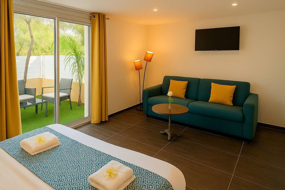 A hotel room with a sofa, a small round table, a TV mounted on the wall, and a sliding glass door leading to an outdoor area with chairs and a table.