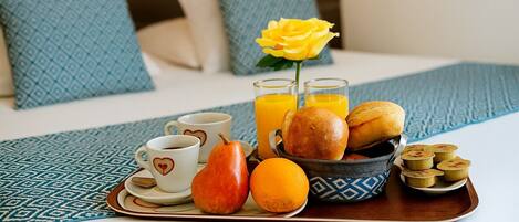 Ein Frühstücksbrett mit Kaffeetassen, Brot, Obst und Saft auf einem Bett mit gemusterten Kissen.