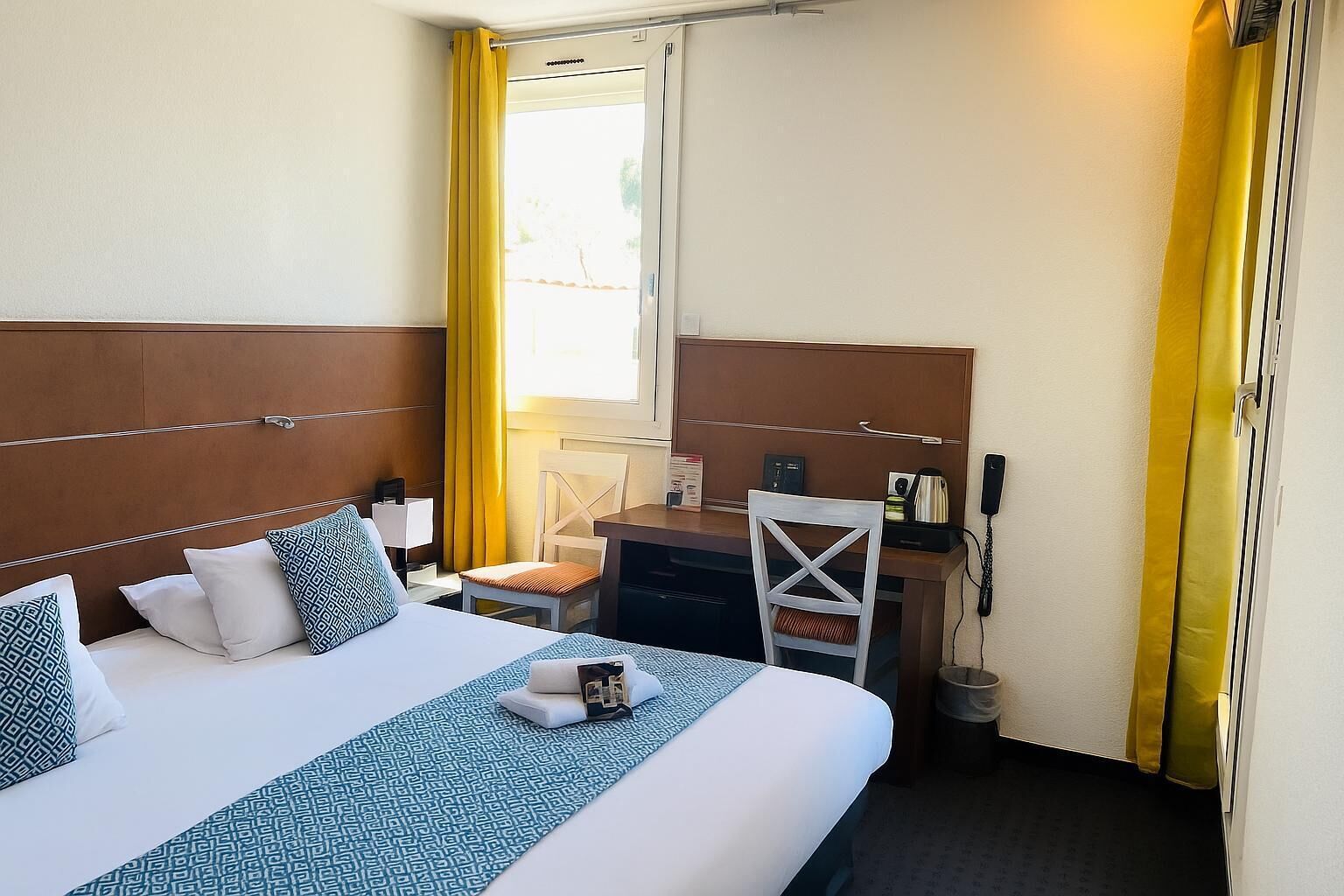 Une chambre d’hôtel avec un lit, un bureau, une chaise et une fenêtre avec des rideaux jaunes.