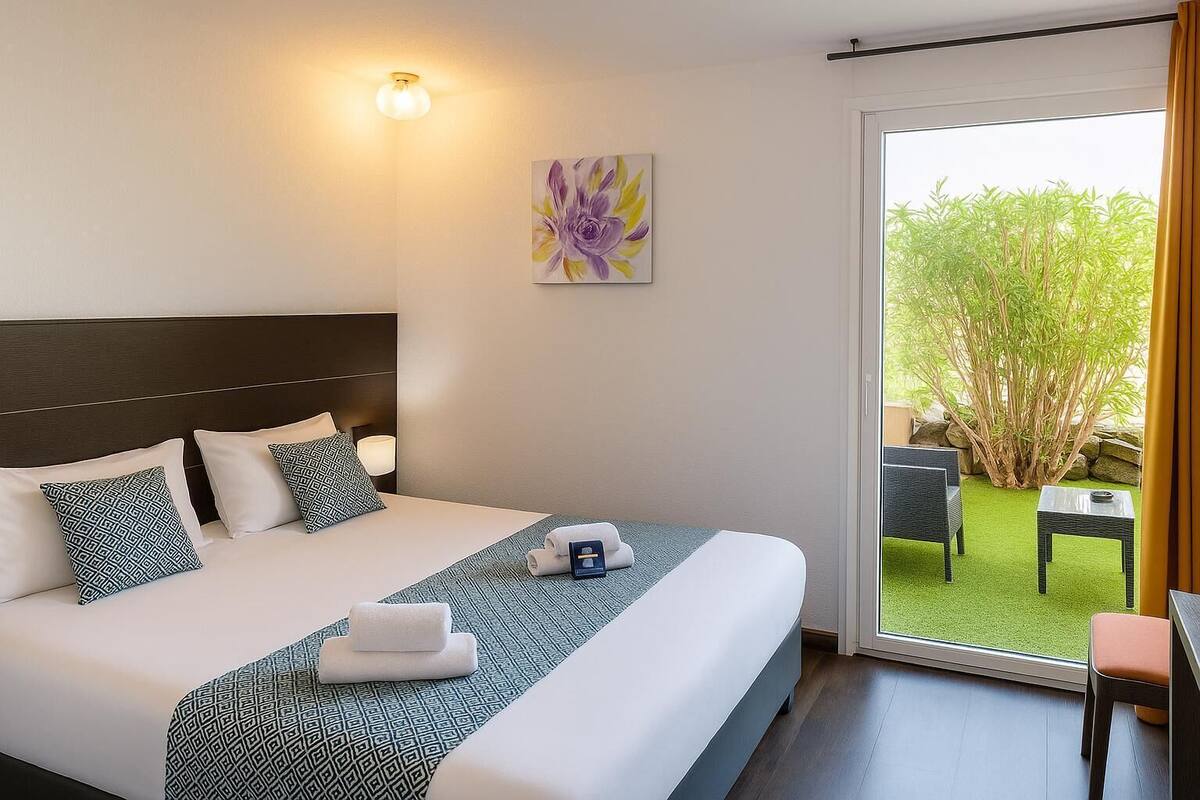 A hotel room with a large bed, a headboard, a painting, and a view of an outdoor area with a table and chairs.