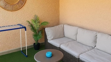 A corner with a wicker sofa, a small round table with a blue object, a blue drying rack, and a potted plant.