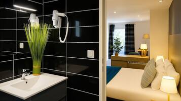 A modern bathroom with a white sink, black tiled walls, and a green plant. It also features a bed with a bedside table, a lamp, and a window with curtains.