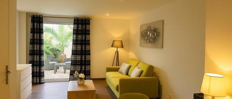 A modern living room with a green sofa, a wooden coffee table, and a lamp. There is a painting on the wall and a view of greenery outside.