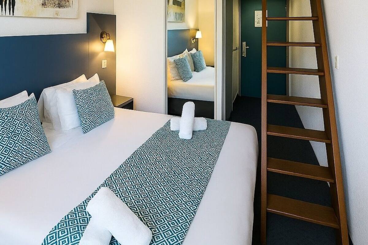 A hotel room with a large bed, a wooden ladder, a mirror, and a painting on the wall.