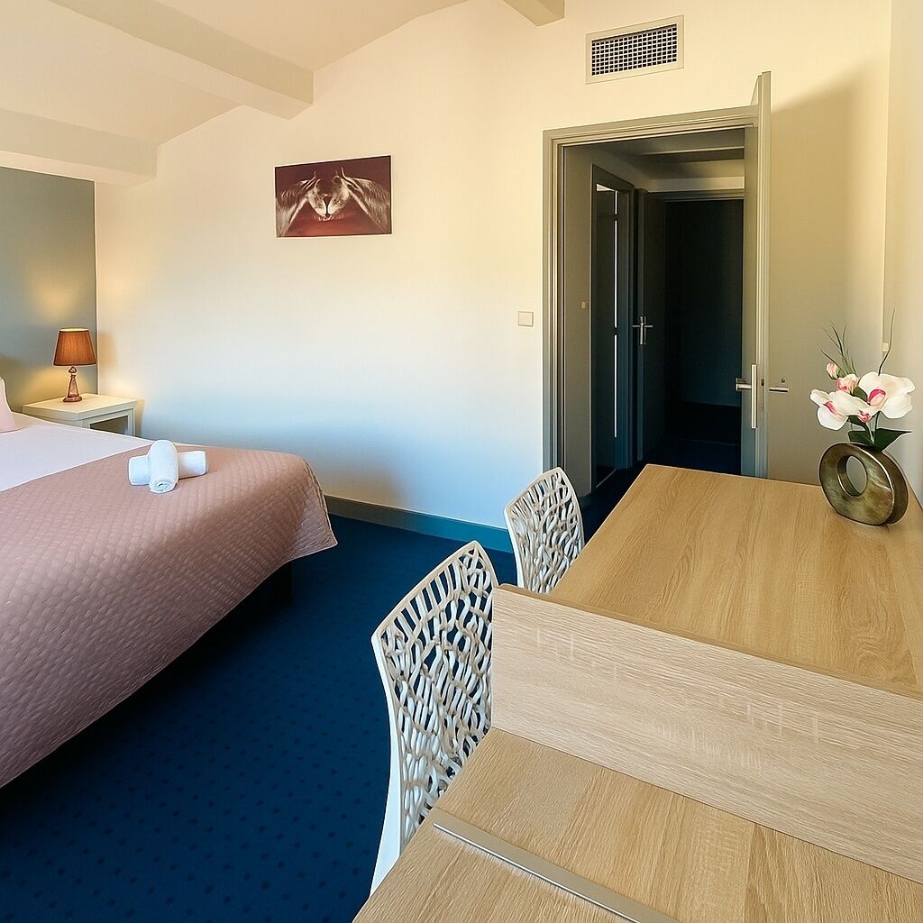 A hotel room with a bed, a desk, and a chair. There is a picture on the wall and a vase with flowers on the desk.