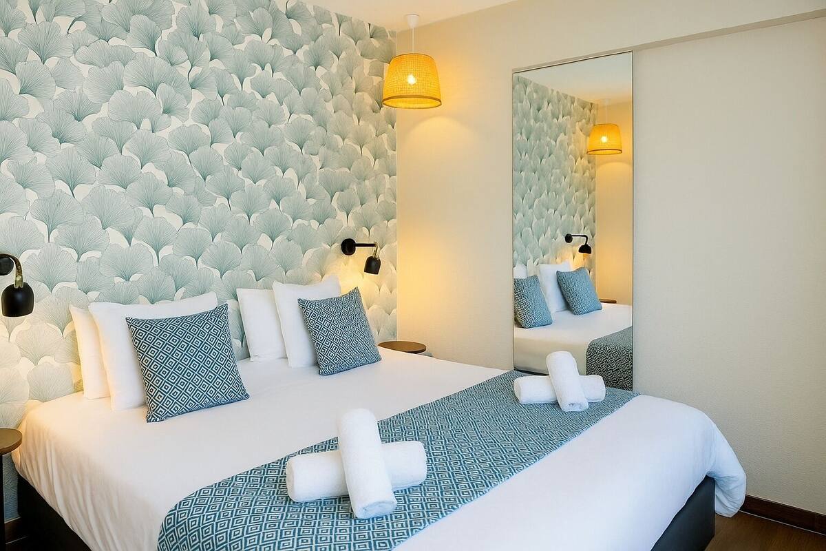 A hotel room with a patterned wallpaper, a bed with white and blue bedding, two wall-mounted lamps, and a large mirror.