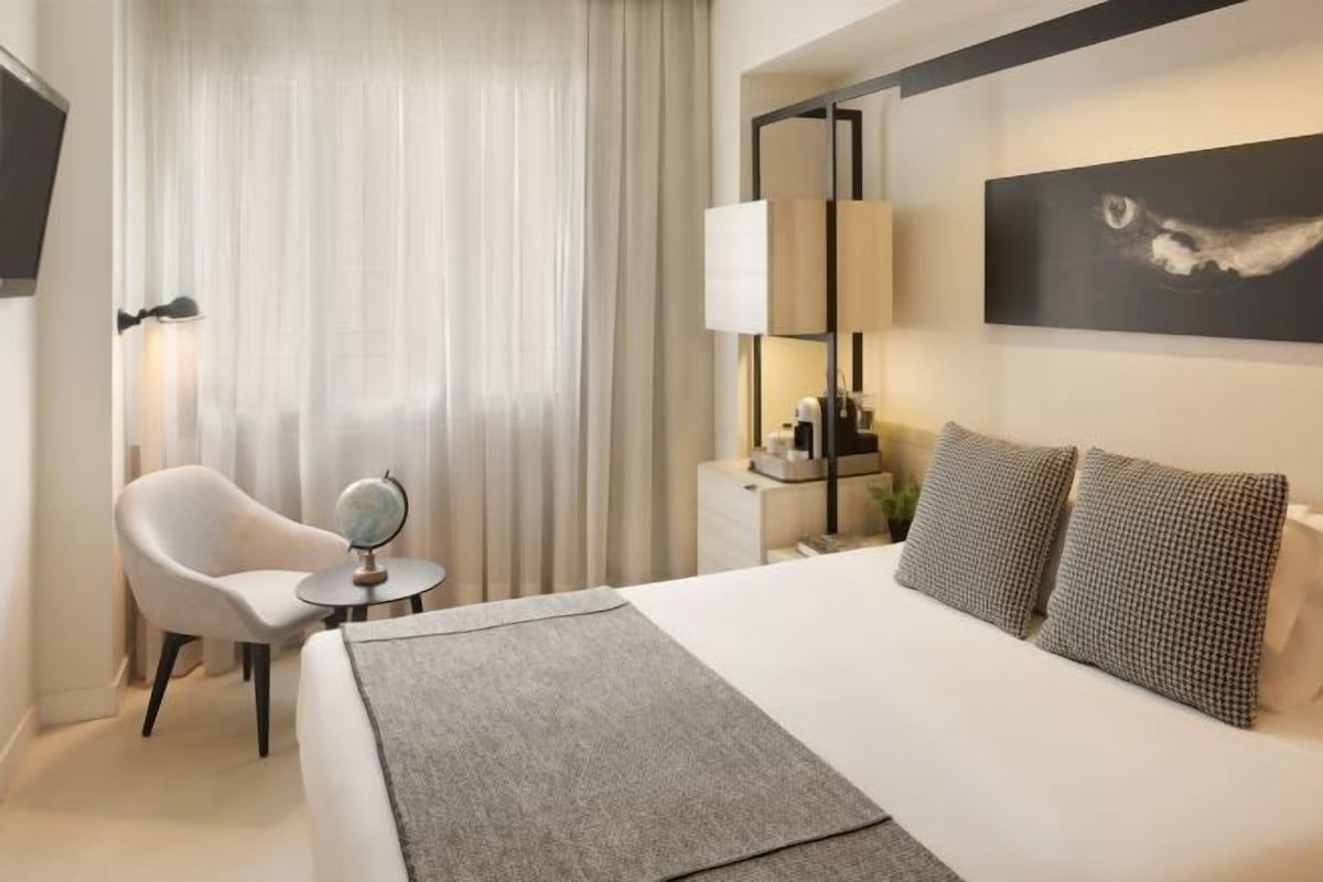 A hotel room with a bed, a chair, a side table, a lamp, a mirror, and a painting on the wall.