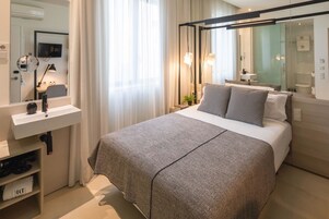 Quarto de hotel moderno com uma cama grande, uma mesinha de cabeceira, um espelho, uma TV e um banheiro com box de vidro.