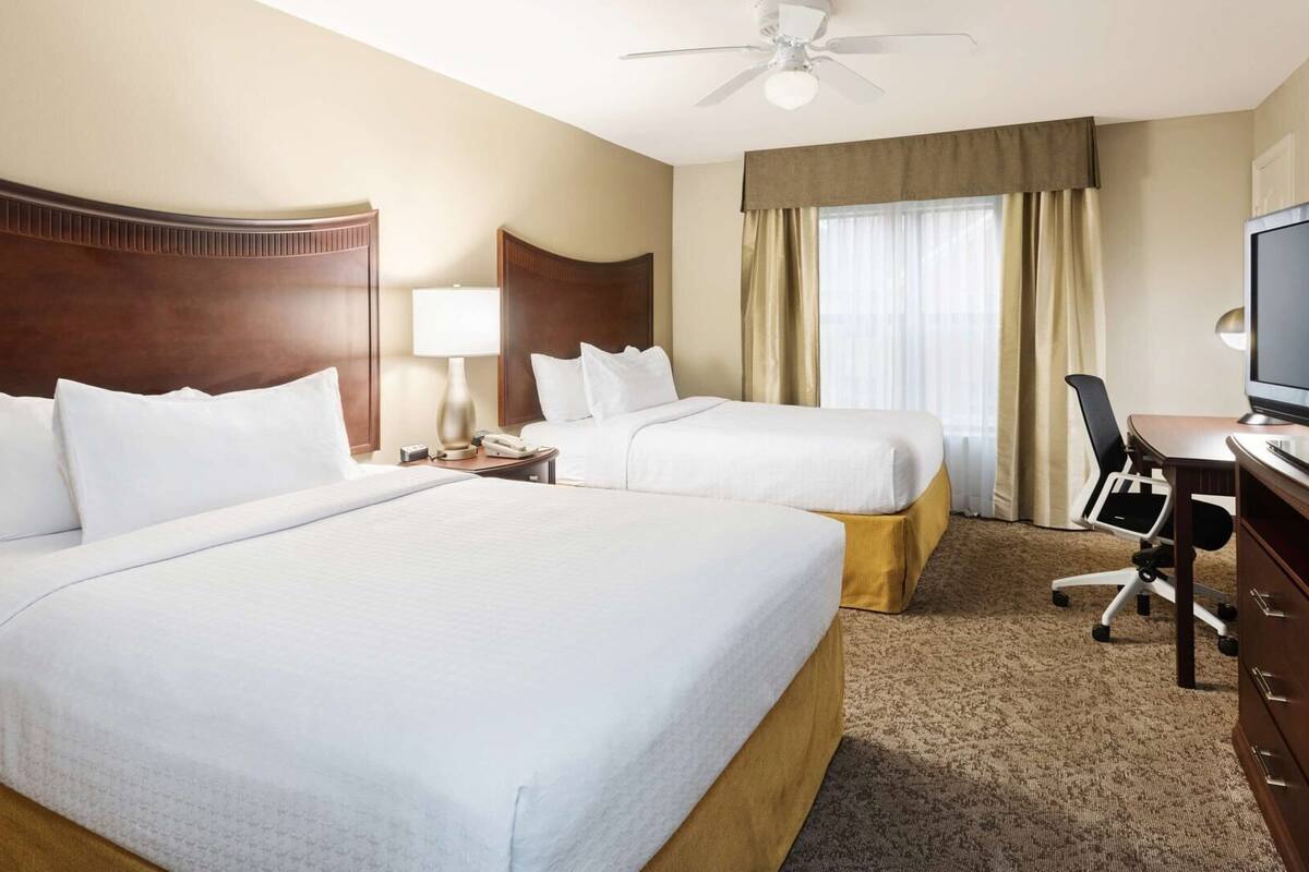 A hotel room with two beds, a television, a desk with a chair, a lamp, and a ceiling fan.