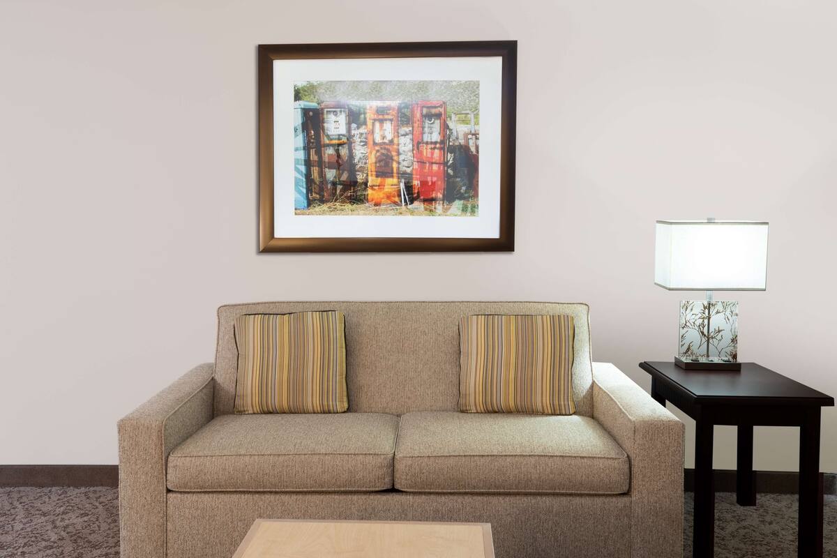 A living room with a beige sofa, two striped pillows, a framed picture on the wall, a small table with a lamp, and a carpeted floor.
