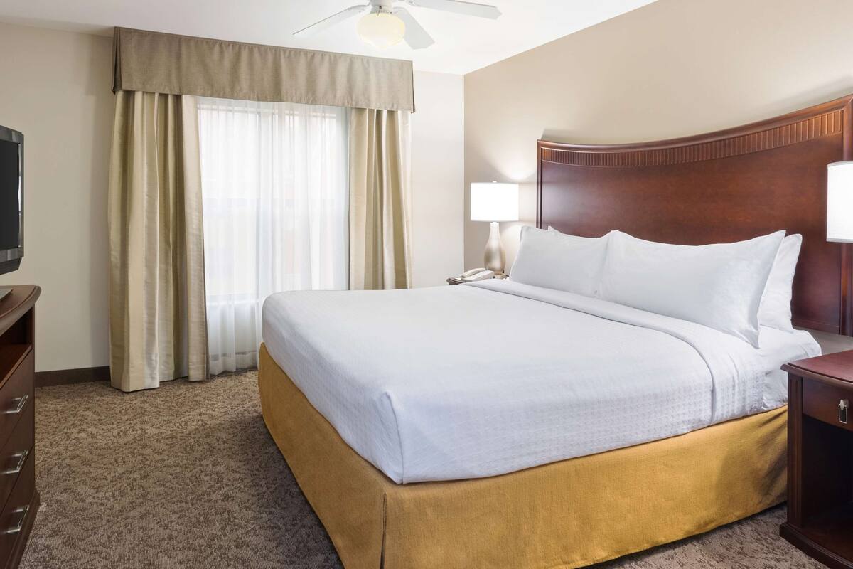 A hotel room with a bed, a television on a wooden dresser, a nightstand with a lamp, and a window with curtains.