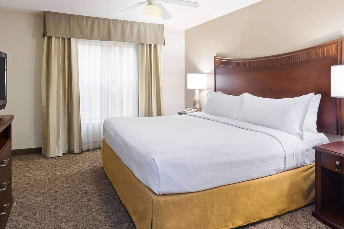 A hotel room with a bed, a television on a dresser, a ceiling fan, and a window with curtains.