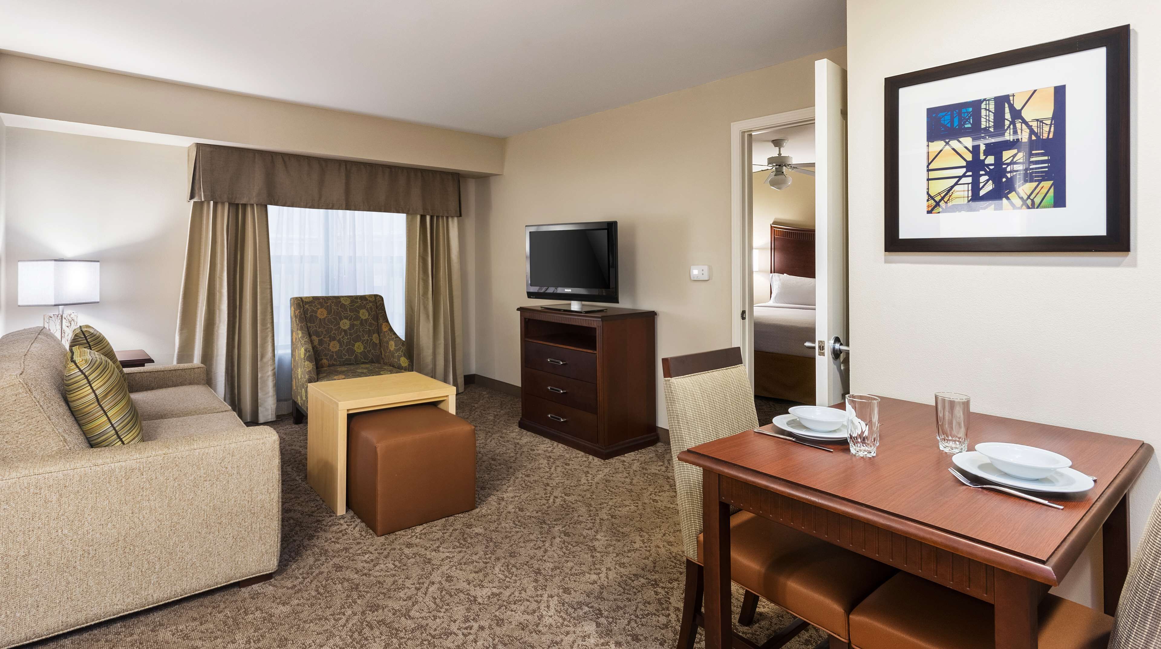 A hotel room with a sofa, a small table with a television, a dining table with a chair, and a bed in the background.