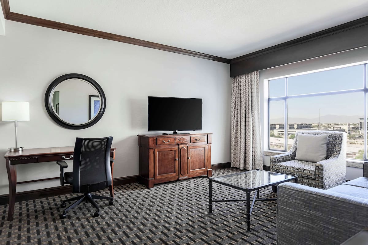 A hotel room with a desk, chair, TV, cabinet, and a view of the city through large windows.