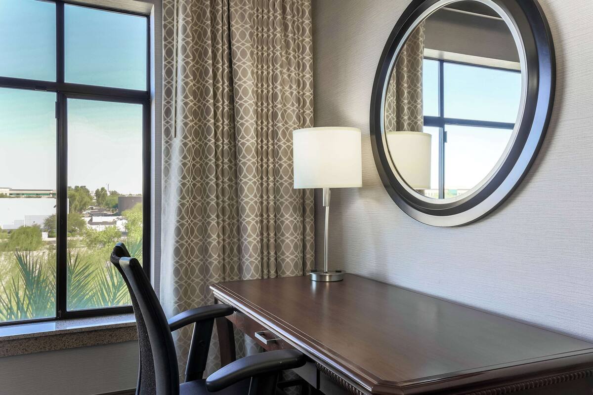 A hotel room with a large window, a round mirror, a table, and a chair.