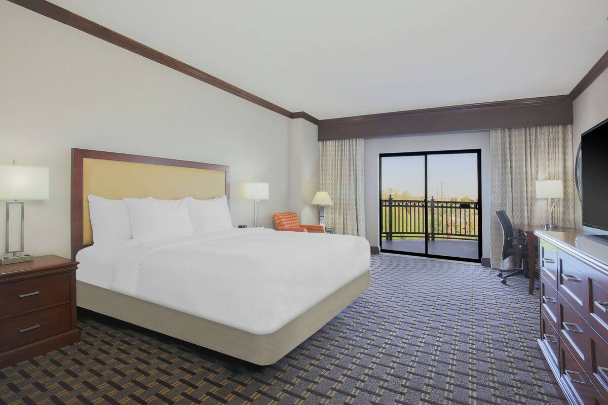 A hotel room with a large bed, two bedside tables with lamps, a desk with a chair, and a view of the outdoors through a sliding glass door.