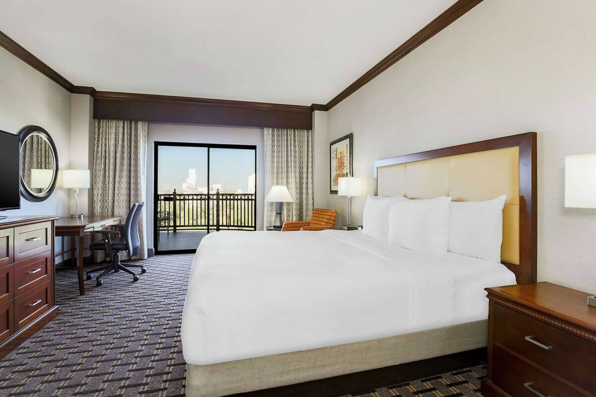A hotel room with a large bed, a desk with a chair, a television, and a balcony with curtains.