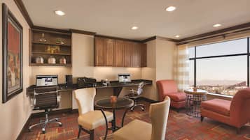 A hotel room with a desk, two chairs, a small table, a bookshelf, and a large window with a city view.