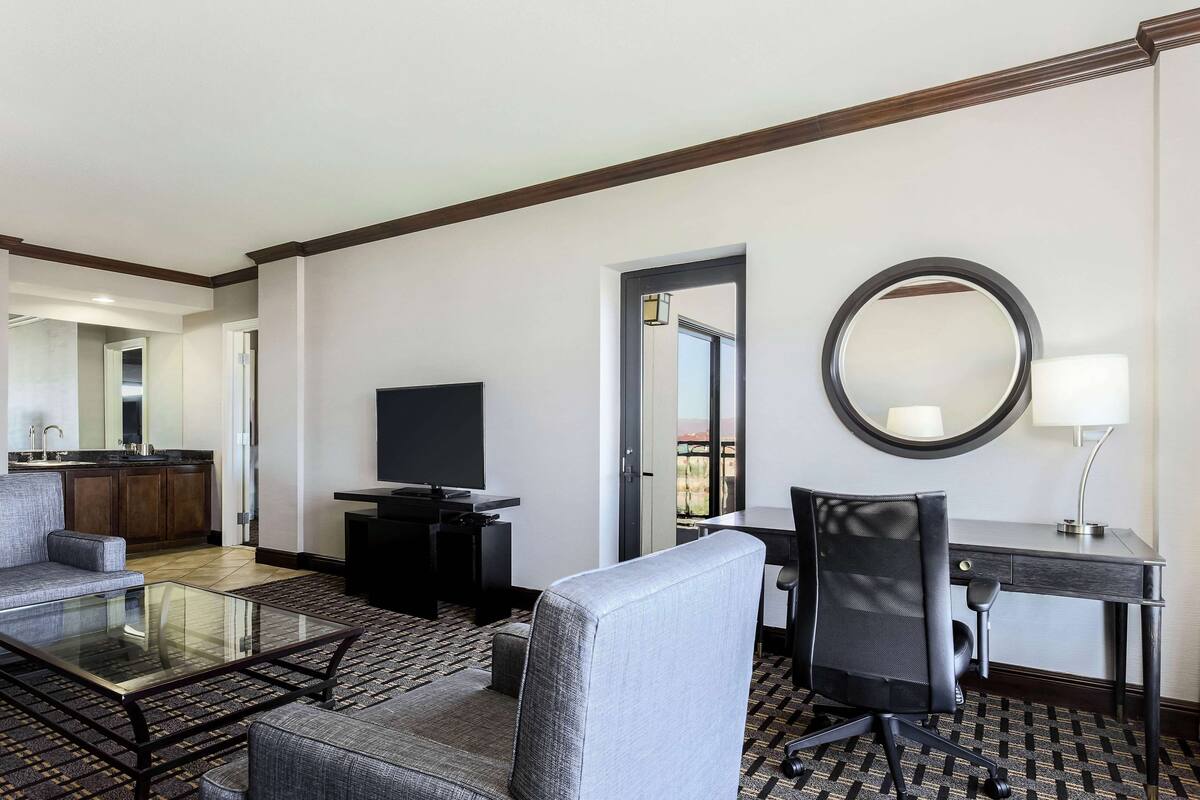 A hotel room with a sofa, a glass coffee table, a desk with a chair, a TV, a mirror, and a lamp.