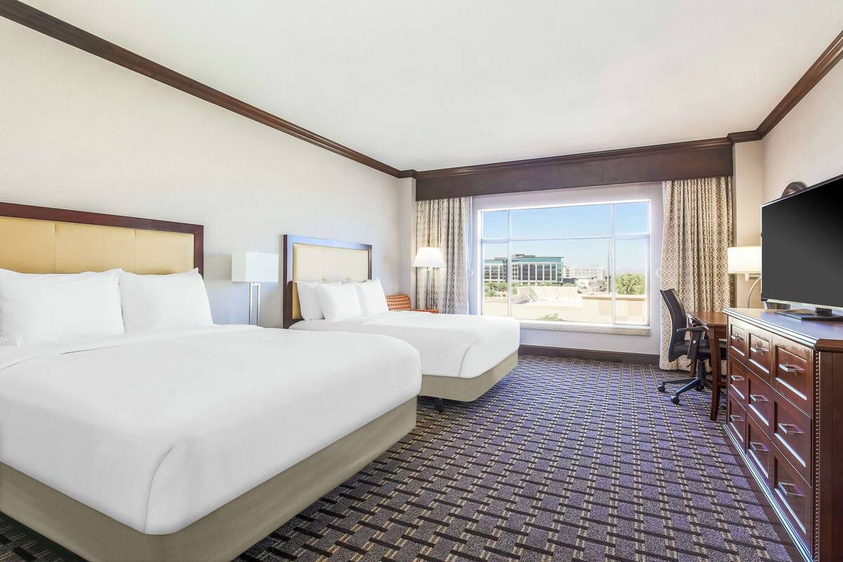 A hotel room with two beds, a desk, a television, and a large window offering a city view.
