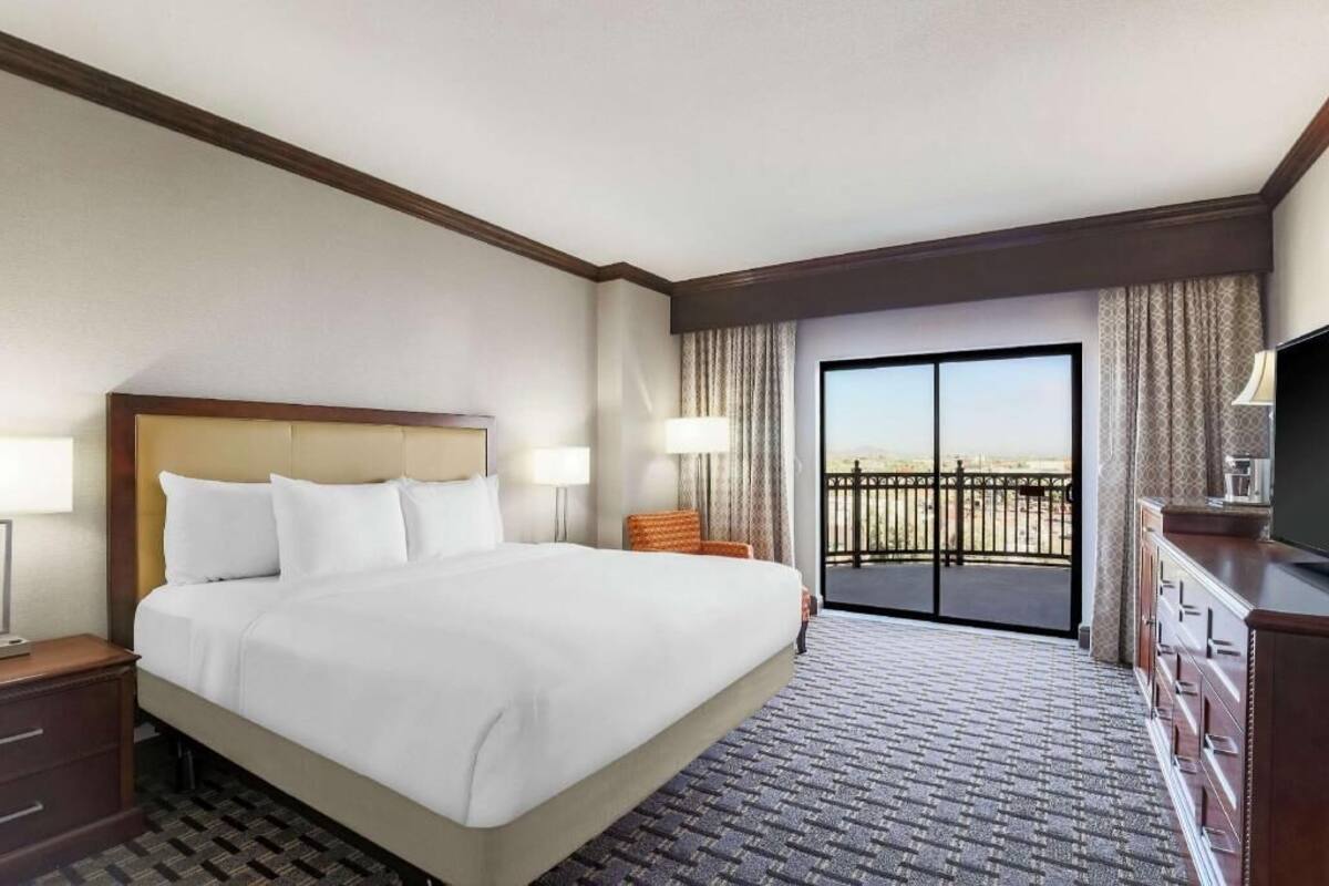 A hotel room with a large bed, a television, a desk with drawers, a chair, and a balcony with a view.
