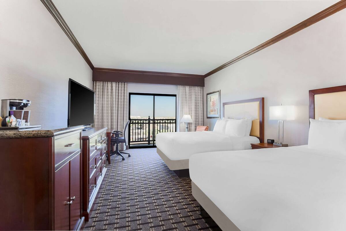 A hotel room with a large bed, a desk with a chair, a dresser, and a TV. There is a balcony with a view.