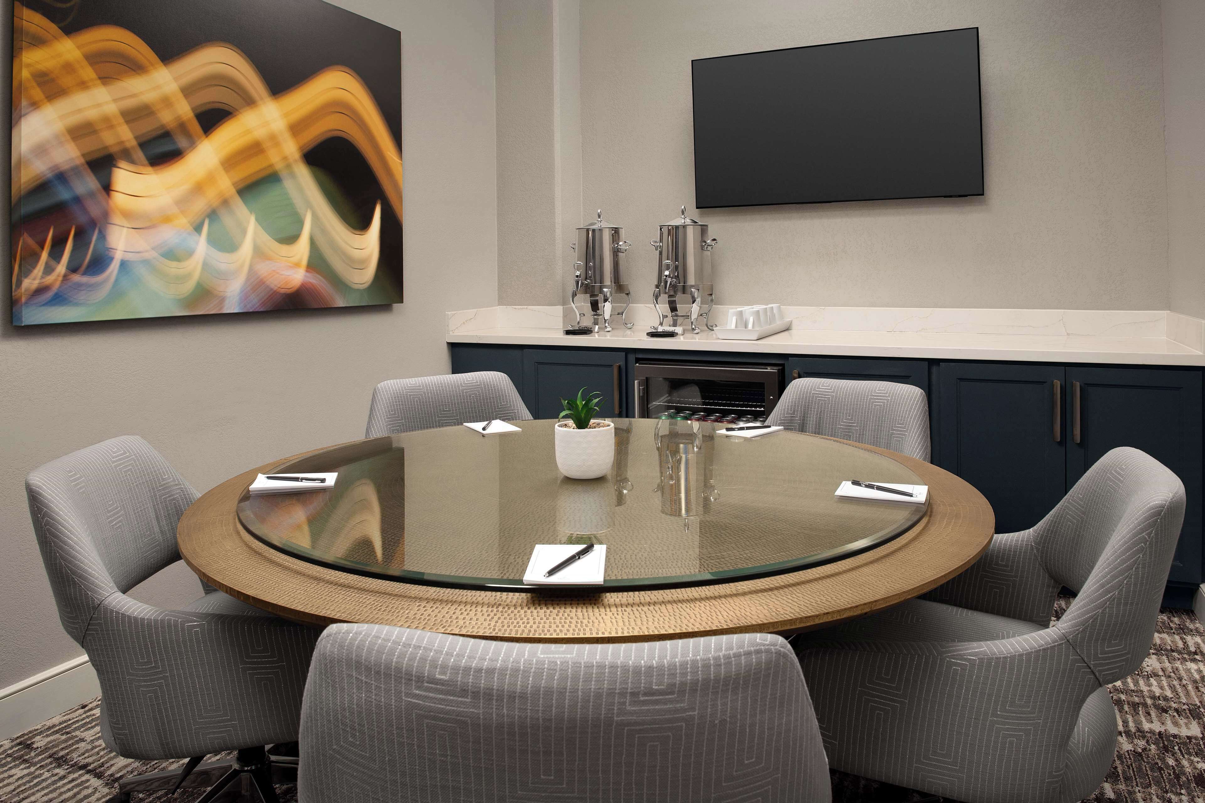 A conference room with a round glass table, six grey upholstered chairs, a coffee station, and a large abstract painting on the wall.