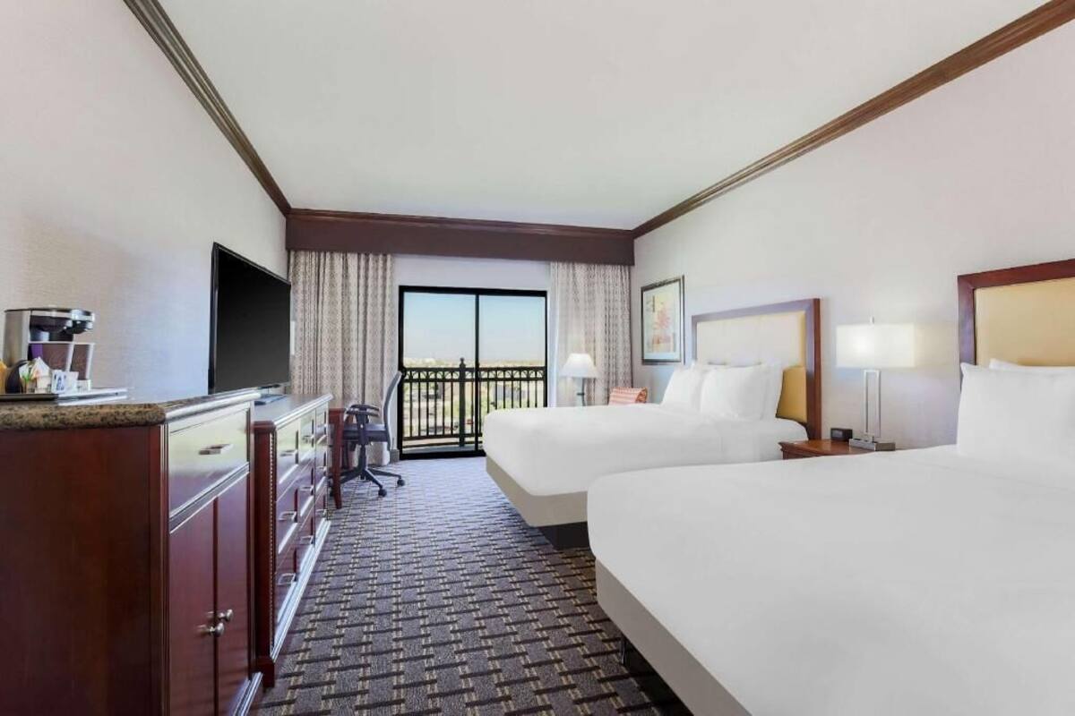 A hotel room with two beds, a desk with a flat-screen TV, a minibar, a desk lamp, and a view of the outdoors through a sliding door.